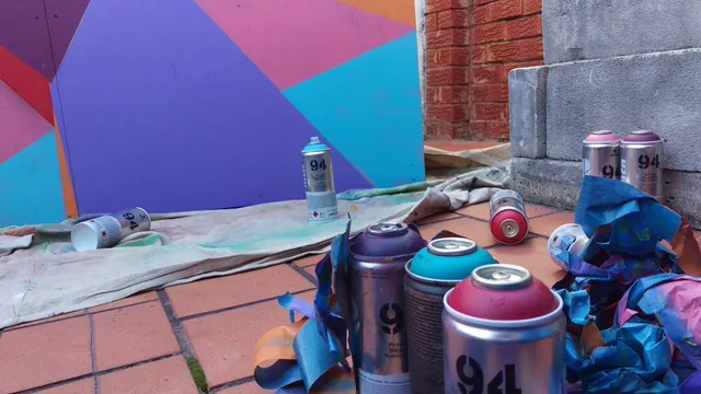 Spray paint cans and crumpled masking tape on a tiled floor in front of a colorful geometric mural in progress