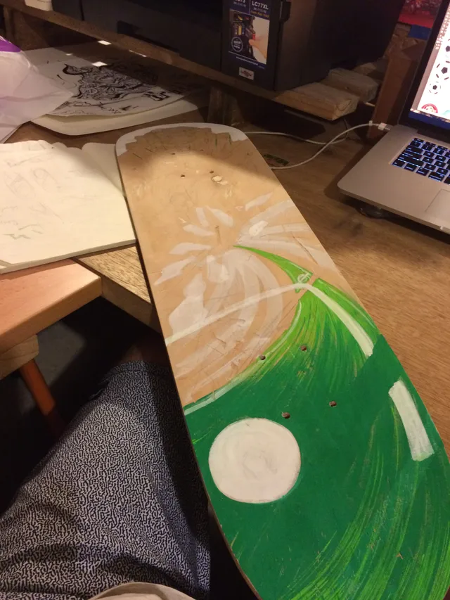 Partially painted wooden skateboard deck on a desk showing swirling green paint and white circular outlines