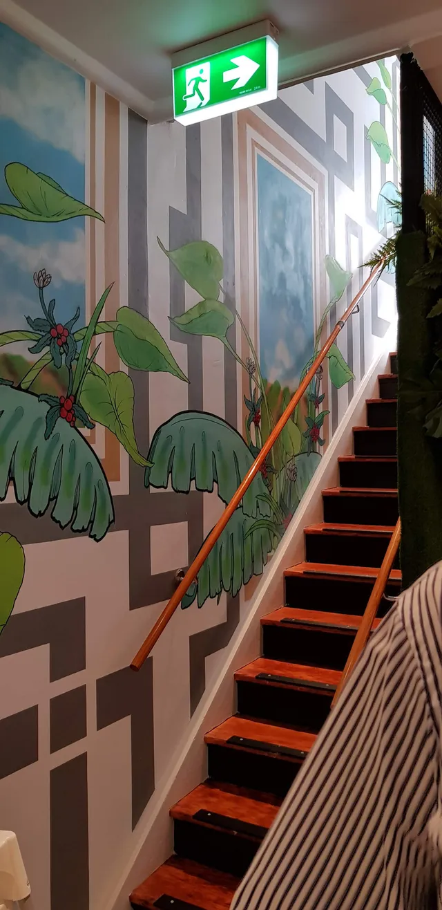 Stairwell mural of tropical leaves, coffee plants, and grey geometric patterns with blue sky window panels