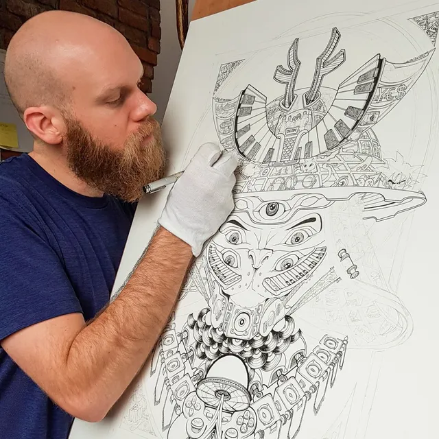 Artist drawing a detailed black ink poster of a symmetrical, multi-eyed totemic face wearing a samurai-style helmet