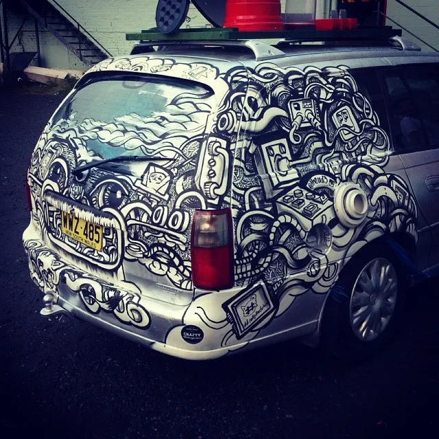Silver car hand-painted with bold black and white line art of tangled cables, electrical plugs, and eyes