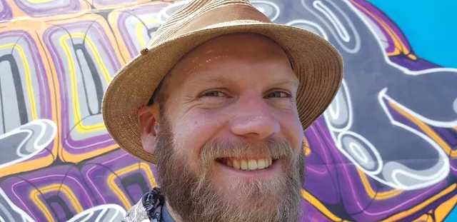 Smiling bearded man in a straw hat in front of a vibrant purple and yellow mural with bold psychedelic linework
