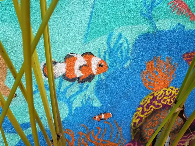 Painted mural on a textured blue wall depicting clownfish and vibrant coral, viewed through green plant stalks