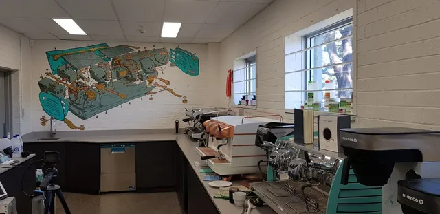 Teal and copper mural of an exploded espresso machine diagram on a white brick wall behind espresso machines