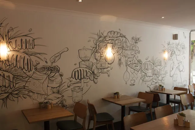 Black line art mural on a cafe wall illustrating coffee beans, abstract brewing machinery, plants and a barista