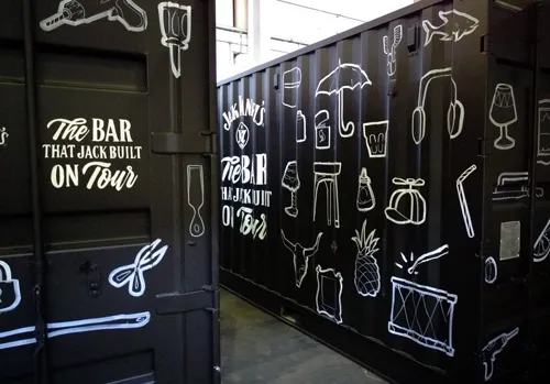 Hand-painted black shipping containers with white line art of bar items and 'The Bar That Jack Built On Tour'