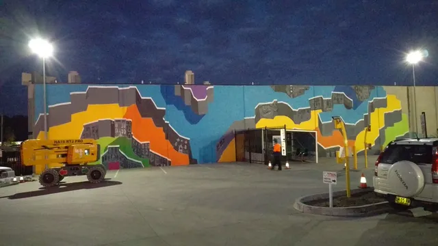 Exterior mural of colorful, abstract geometric structures on a long wall at night, with a yellow lift parked in front
