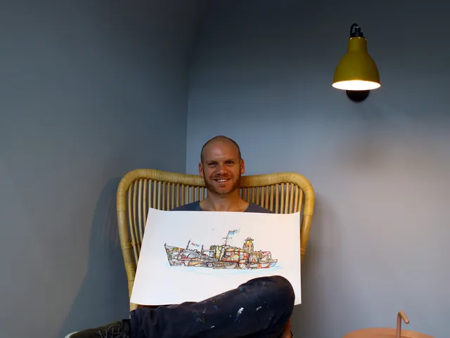 Smiling man in a wicker chair holding a large ink and color illustration of a detailed ship
