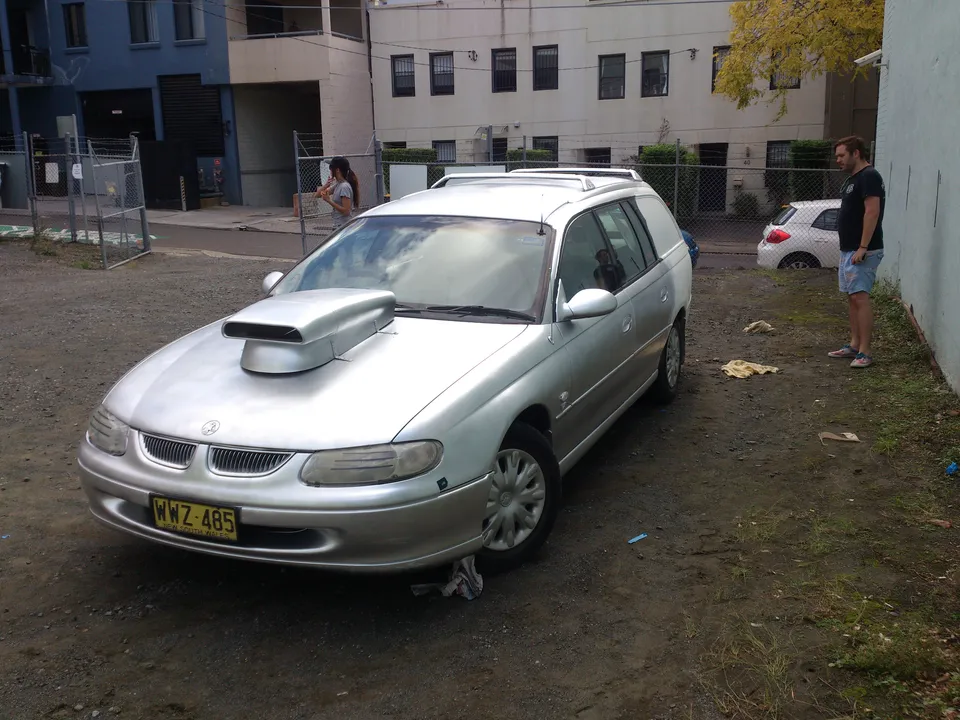 Plain silver station wagon with a large hood scoop parked in a dirt lot
