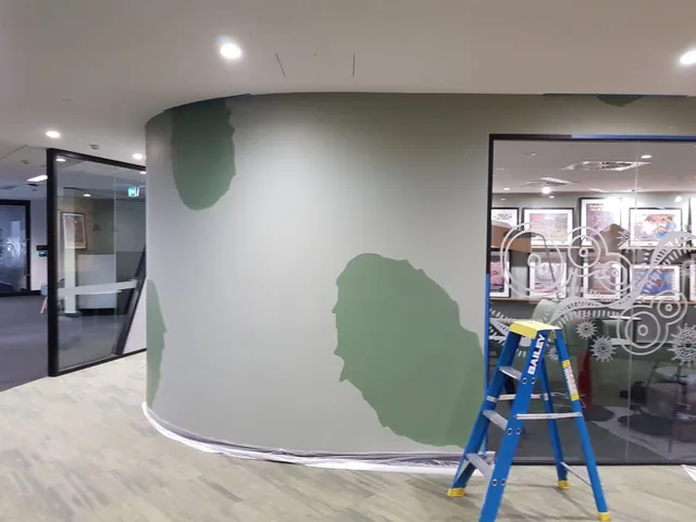 Mural in progress on a curved office wall showing large patches of green paint over a grey base and a blue stepladder
