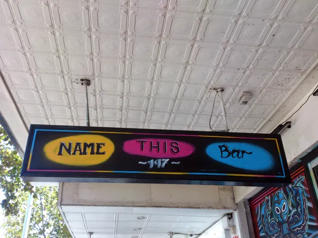 Hand-painted hanging sign reading Name This Bar ~197~ over yellow, pink, and blue spray-painted ovals