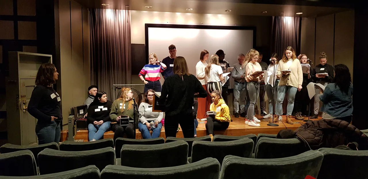 People on a stage holding papers during a rehearsal, viewed from empty theater seats