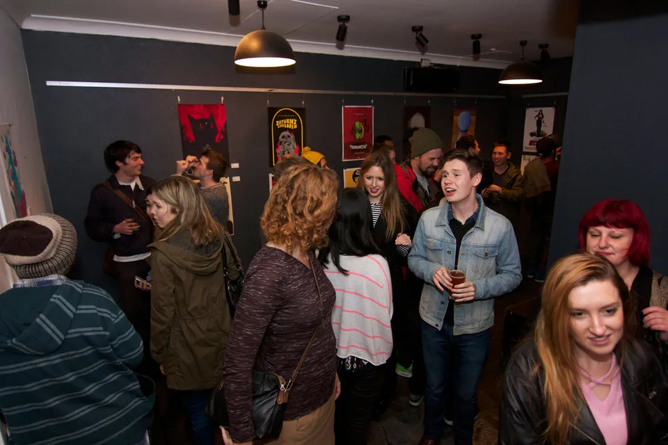 Crowd of people mingling at a gallery exhibition with colorful posters hanging on dark walls in the background
