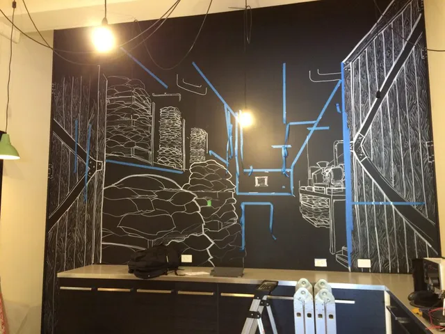 In-progress white line mural on a black wall depicting stacked coffee sacks and barn doors, marked with blue tape