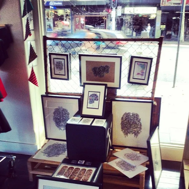 Framed intricate illustrations and prints displayed on pallets and a wire mesh screen in a shop window