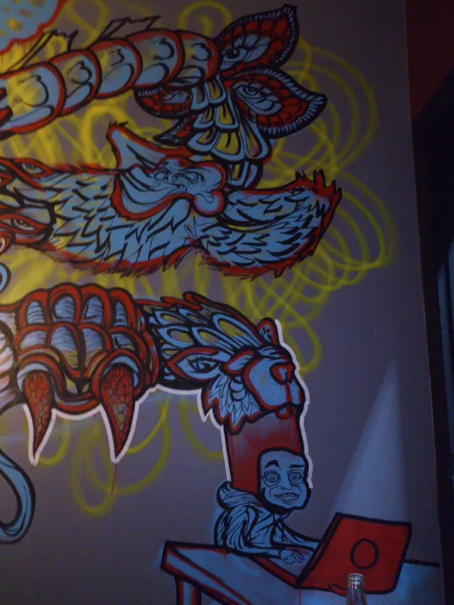 Wall mural featuring a stylized figure at a red laptop below towering psychedelic animal masks and a giant red claw
