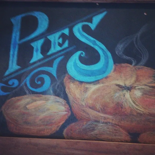 Chalk drawing on a blackboard showing the word Pies in stylized blue lettering above two steaming baked pies