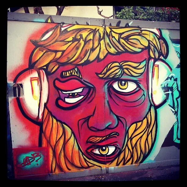 Mural of a stylized red face with a yellow beard, headphones, and a third eye on its chin