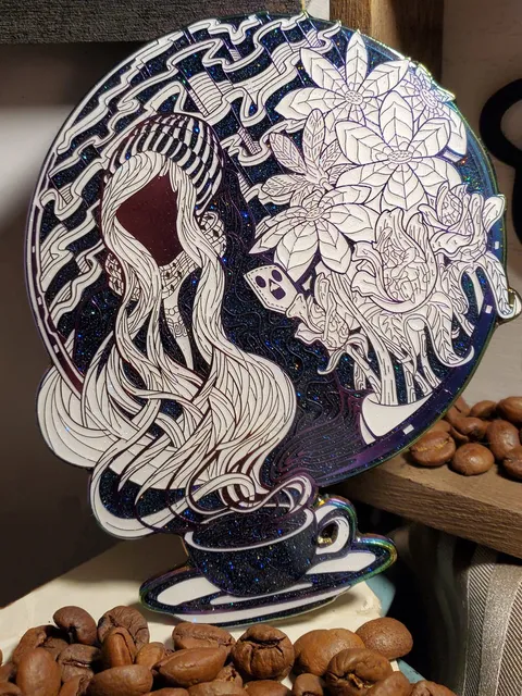 Glittery enamel pin of a faceless woman in a beanie rising like steam from a coffee cup, placed among coffee beans