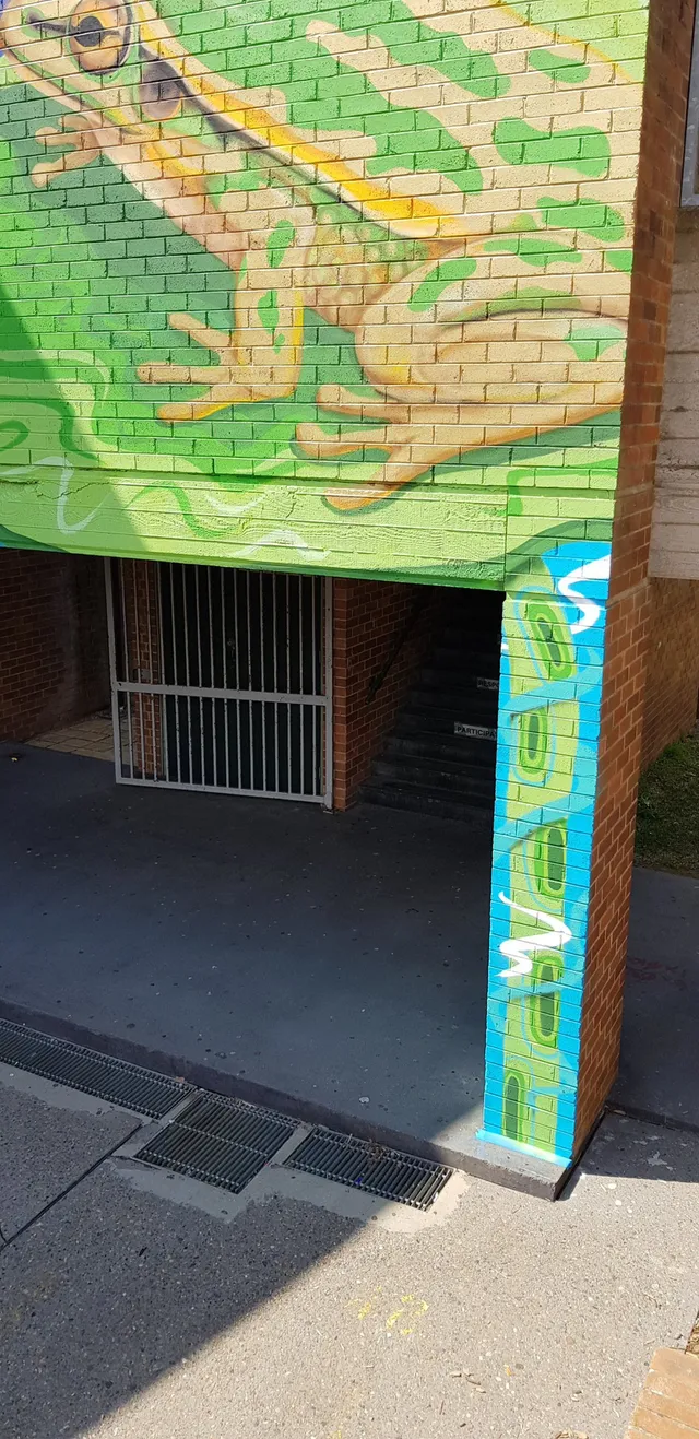 Green and yellow frog mural on a brick wall over a gated stairwell beside a blue patterned pillar