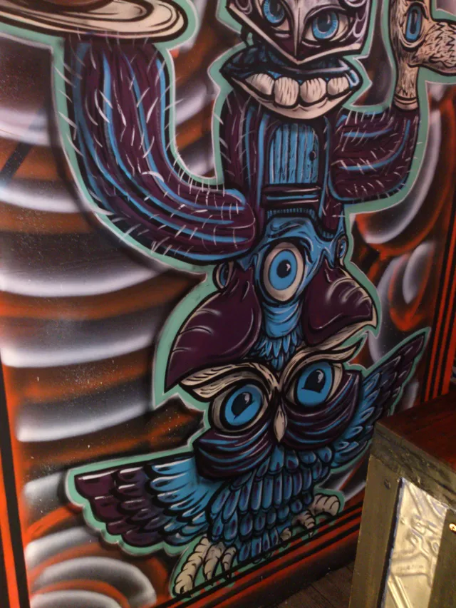 Mural of a psychedelic blue and purple totem pole featuring an owl, a central eye, and a grinning masked figure