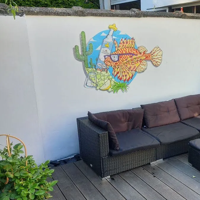 Mural on a white wall featuring a lionfish in sunglasses, a tequila bottle, and a cactus above a black wicker sofa
