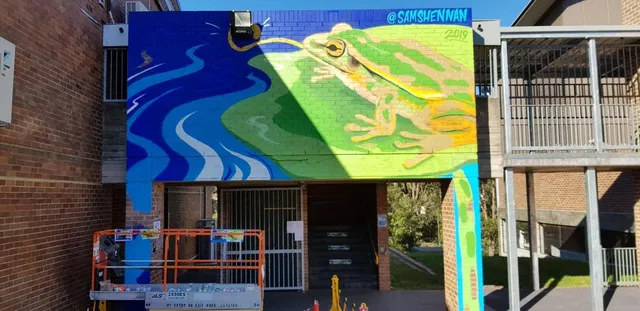 Mural on a brick wall of a large green frog beside a blue river catching a bug with its extended tongue