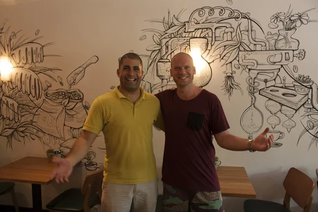 Two smiling men posing in front of an indoor wall mural featuring black line art of stylized coffee brewing equipment