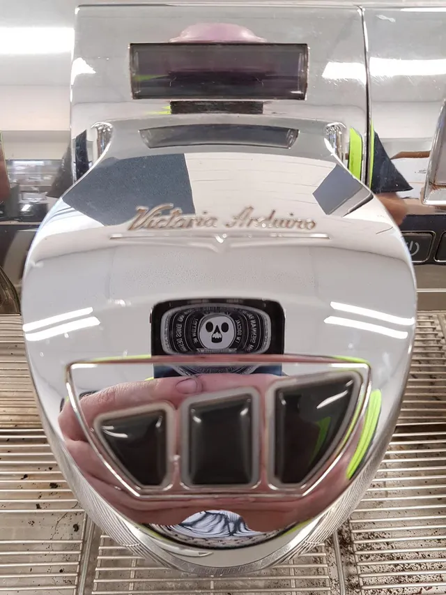 Chrome Victoria Arduino espresso machine reflecting a hand holding a phone with a skull logo