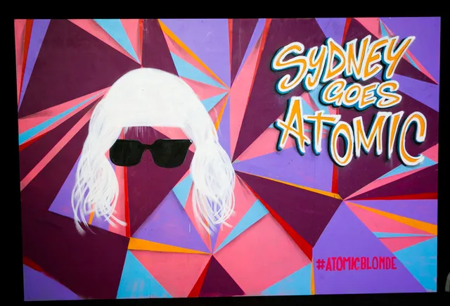 Mural of a blonde wig and sunglasses on a pink and purple geometric background reading SYDNEY GOES ATOMIC