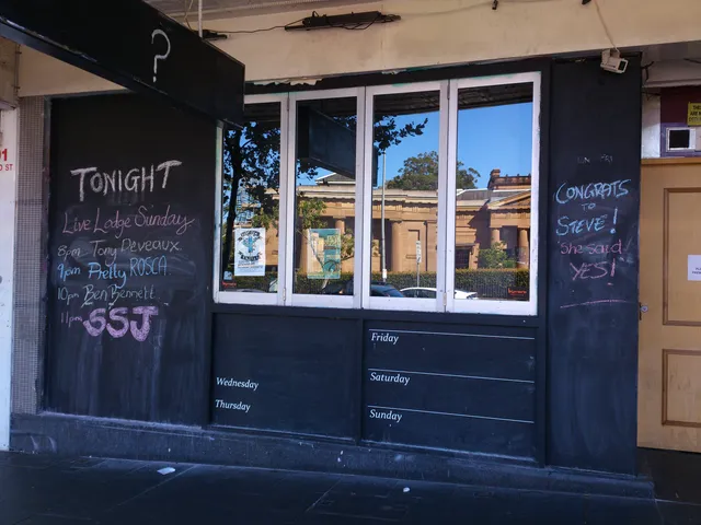 Chalk hand-lettering on a black bar exterior detailing a gig lineup below a hanging sign with a question mark