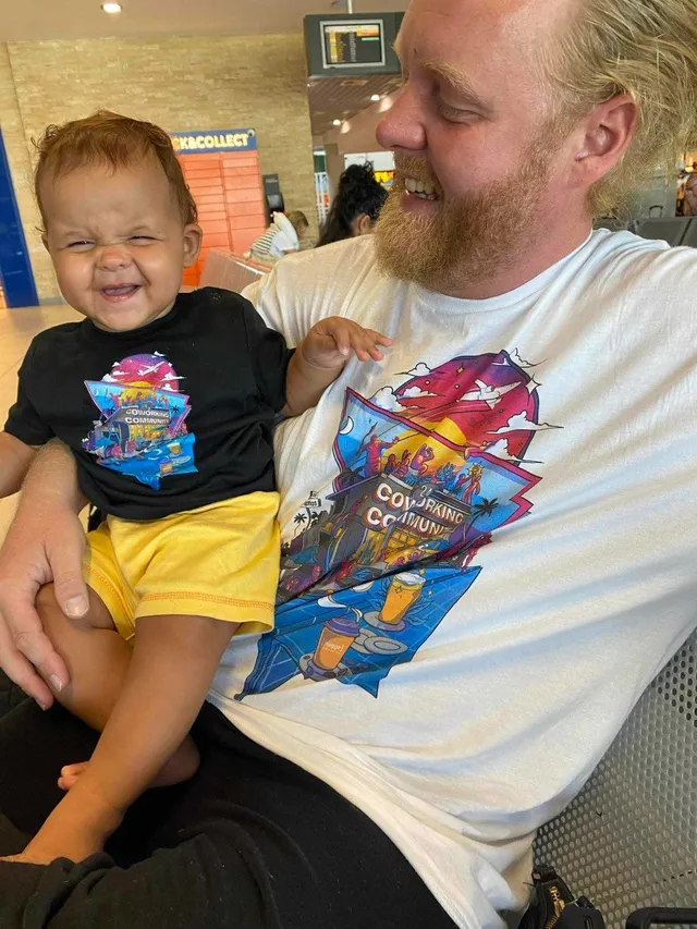 Smiling man and baby wearing t-shirts printed with a vibrant graphic illustration reading Coworking Community
