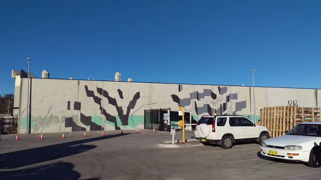 Work-in-progress mural on an exterior concrete wall showing bold black and grey geometric shapes and faint linework