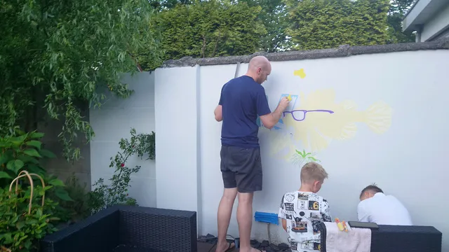Artist painting the early stages of a lionfish mural on a white outdoor wall with two boys nearby