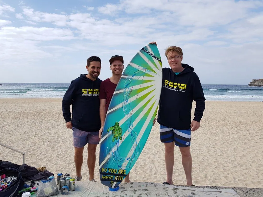 Three men on a beach holding a painted surfboard with a blue and green sunburst, a sea turtle, and text