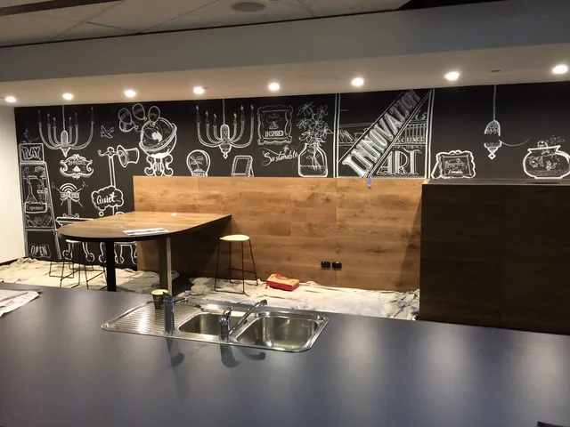 White line-art mural on a black office kitchen wall featuring a globe, chandeliers, and typography