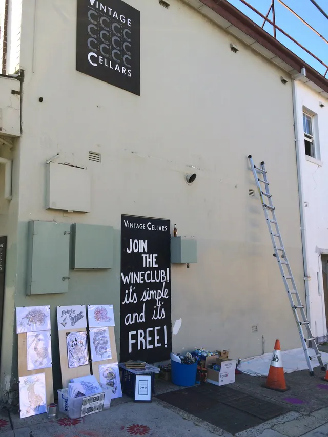 Blank exterior wall prepared for a mural, showing a ladder, paint supplies, and reference sketches on leaning boards