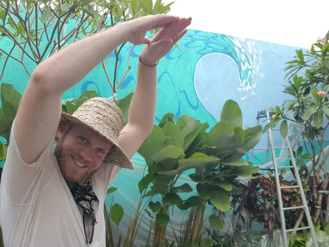 Man in a straw hat posing with raised arms in front of a bright blue wave mural and tropical plants