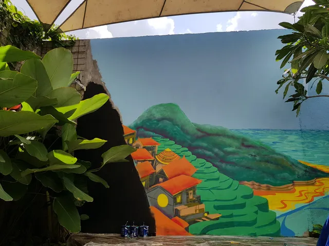 Partially completed outdoor mural depicting green terraced hills, orange-roofed houses, and the ocean