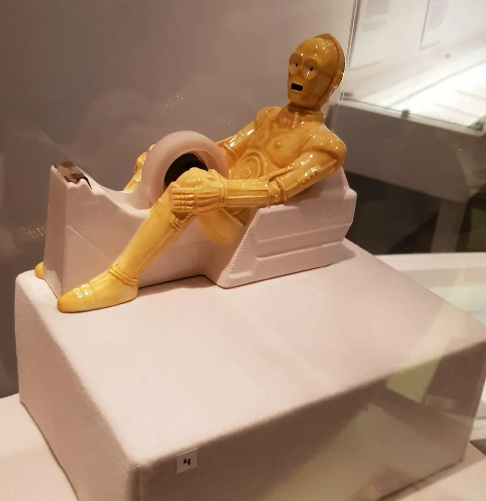 Glossy tape dispenser shaped like C-3PO sitting in a white chair with the tape roll between his legs