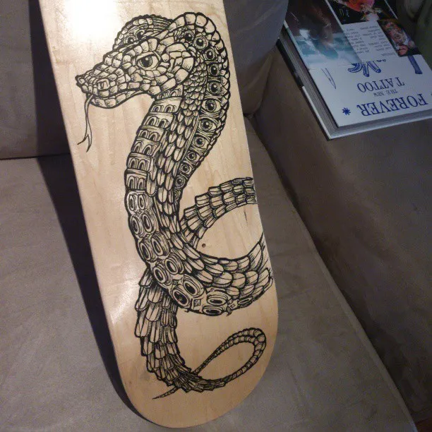 Black ink drawing of a coiled, intricately patterned snake on a wooden skateboard deck
