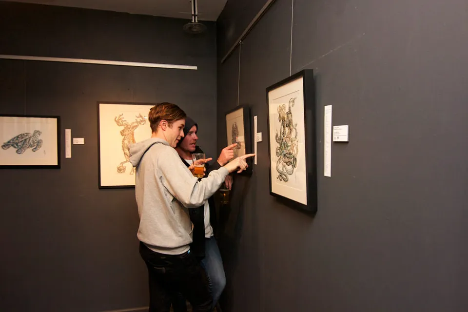 Two gallery attendees pointing at framed ink drawings of animals hanging on a dark grey wall
