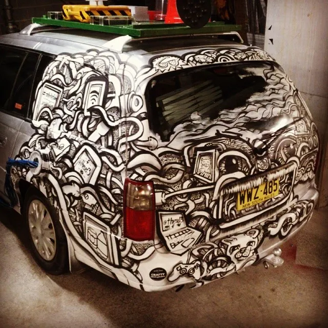 Black and white hand-painted mural on a silver station wagon featuring tangled cables, electronics, and a cat face