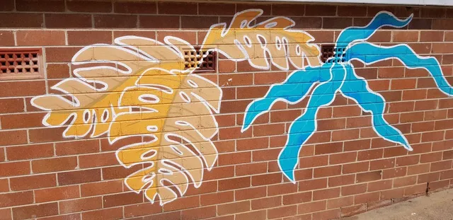 Mural on a brick wall depicting stylized yellow monstera leaves and a wavy blue palm frond outlined in white