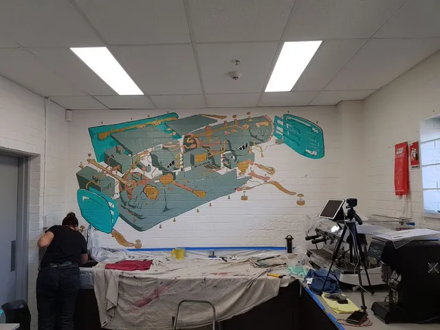 In-progress mural on a white brick wall depicting an exploded diagram of an espresso machine in teal and gold