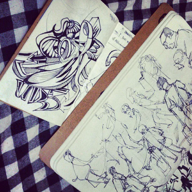Two open sketchbooks on a checkered cloth showing a bold abstract ink drawing and loose figure sketches