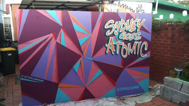 Painted mural panel with abstract purple, pink, and blue geometric shapes and text reading SYDNEY GOES ATOMIC