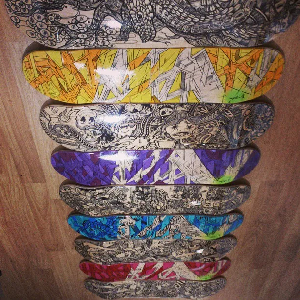 Nine hand-painted skateboard decks featuring intricate black line drawings and bold colorful patterns on a wooden floor