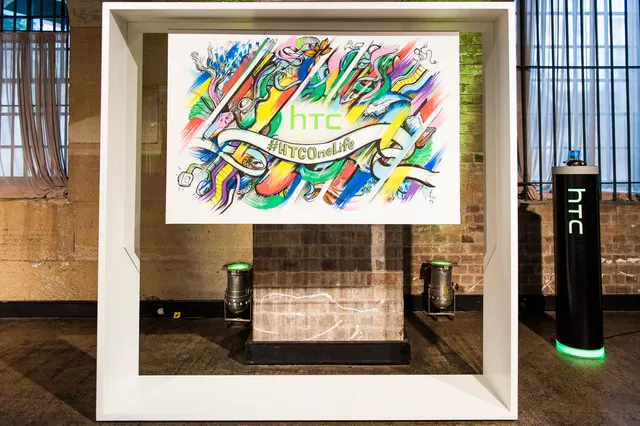 Framed canvas displaying a colorful painting of abstract shapes, the HTC logo, and a #HTCOneLife banner