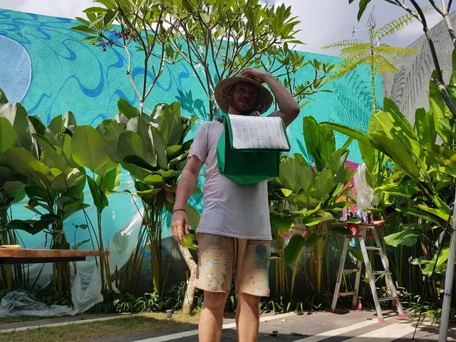 Man in a straw hat and paint-splattered shorts standing before an unfinished blue mural in a tropical garden